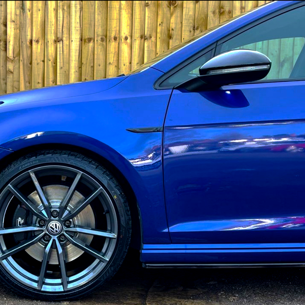 A blue car with a shiny Volkswagen logo on the wheel stands still, next to a wooden fence, reflecting light on its glossy surface.