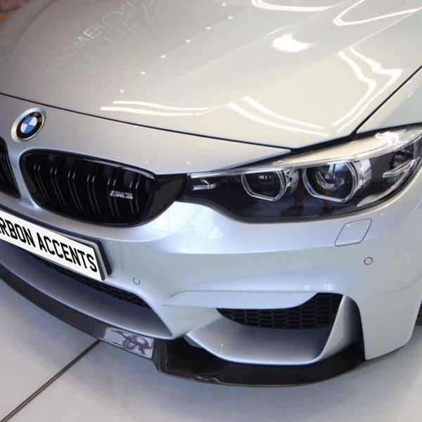 A silver BMW M3 car is parked indoors, showcasing its front grille and headlights. The number plate reads "CARBON ACCENTS." The background features a brick wall.