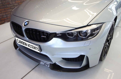 A silver BMW M3 car is parked indoors, showcasing its front grille and headlights. The number plate reads "CARBON ACCENTS." The background features a brick wall.