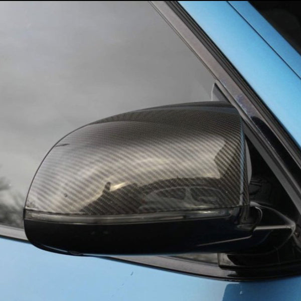 A carbon fiber car side mirror reflects its surroundings, attached to a light blue vehicle, set against a cloudy sky in the background.