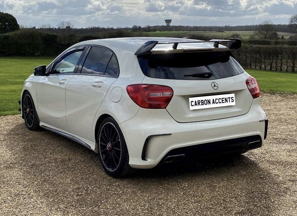 A white hatchback car parked on a gravel driveway, featuring a prominent rear spoiler. The background shows a grassy field and cloudy sky. Text on the license plate reads, "CARBON ACCENTS."