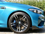 A blue car is parked on gravel, showing its front wheel and headlight. The wheel features a five-spoke design and a visible brake disc. Behind, green shrubs line the background.
