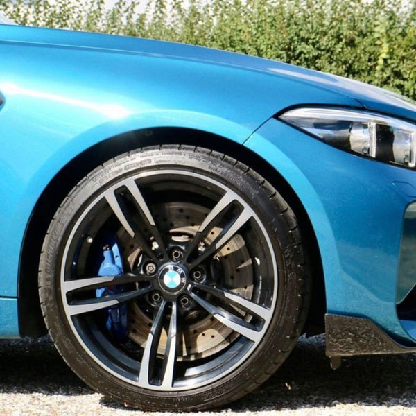 A blue car is parked on gravel, showing its front wheel and headlight. The wheel features a five-spoke design and a visible brake disc. Behind, green shrubs line the background.