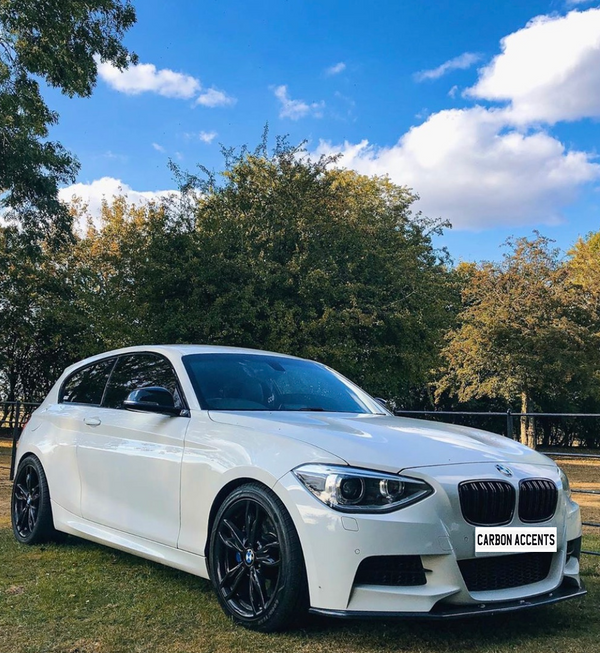 A white car with black rims is parked on grass, surrounded by trees under a blue sky with clouds. Text on the license plate reads "CARBON ACCENTS."