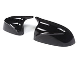 Two glossy black car mirror covers are lying on a plain white background. The covers have sleek aerodynamic designs, reflecting light prominently on their curved surfaces.