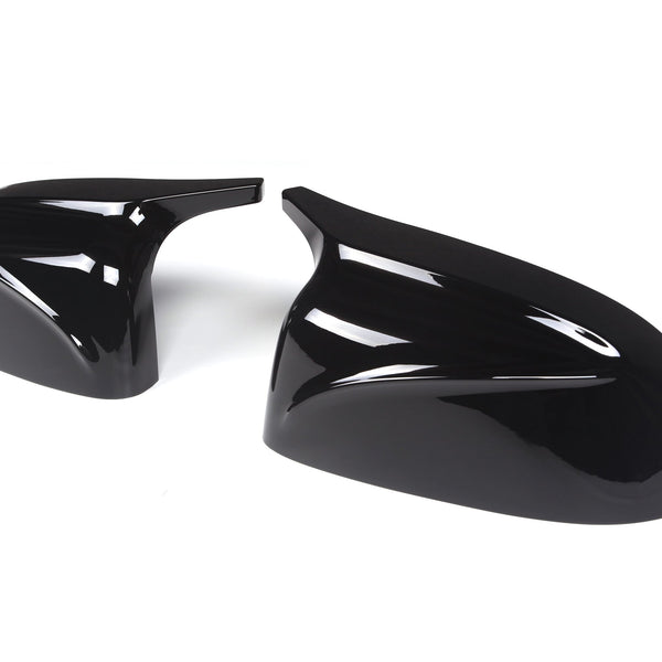 Two glossy black car mirror covers are lying on a plain white background. The covers have sleek aerodynamic designs, reflecting light prominently on their curved surfaces.
