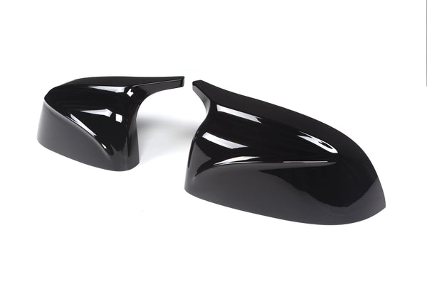 Two glossy black car mirror covers are lying on a plain white background. The covers have sleek aerodynamic designs, reflecting light prominently on their curved surfaces.