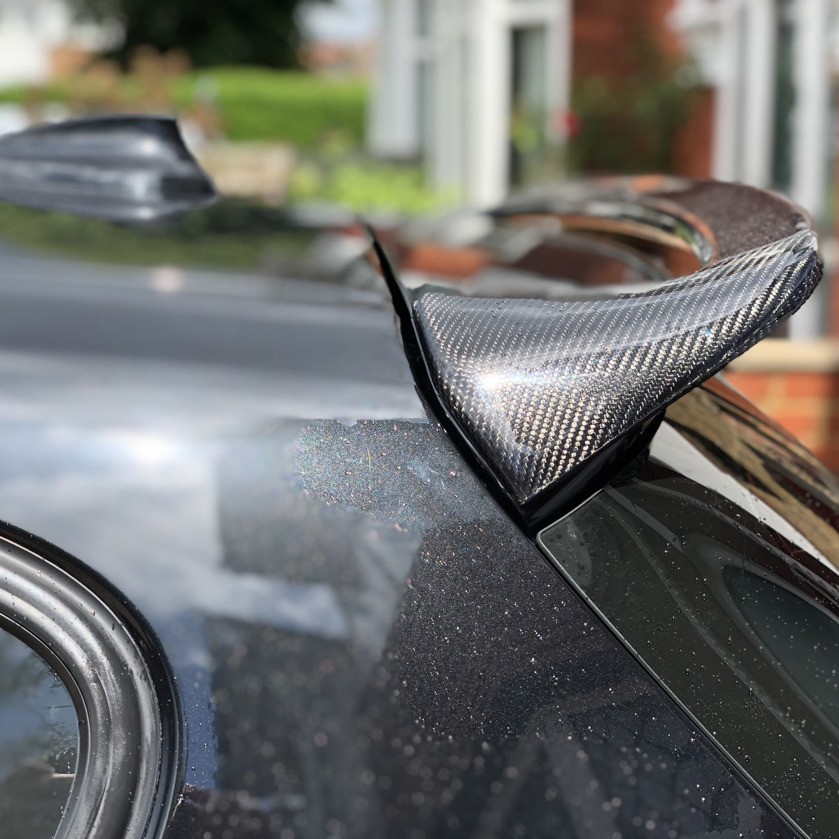 A carbon fiber spoiler is attached to a car's rear in a residential area, reflecting sunlight. A window edge and blurred buildings are visible in the background.