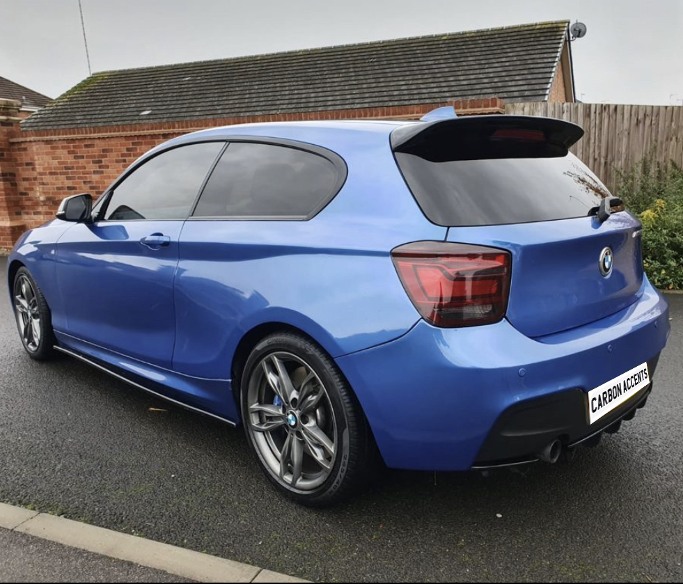 A blue hatchback car is parked on a wet road next to a brick wall. The license plate reads "CARBON ACCENTS," and the environment suggests a residential area.