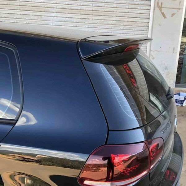 A black car is parked with its rear hatch and spoiler facing a closed metal shutter. The context includes a reflection of an outdoor scene in the car's glossy surface.