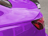 A vibrant purple car features a sleek, tinted taillight and a glossy black spoiler. It is parked on an asphalt street, reflecting nearby buildings.