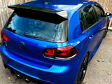 A blue hatchback car is parked on a pavement, featuring a spoiler and dual exhausts. It’s surrounded by wooden fencing and greenery. The Volkswagen logo is visible on the back.