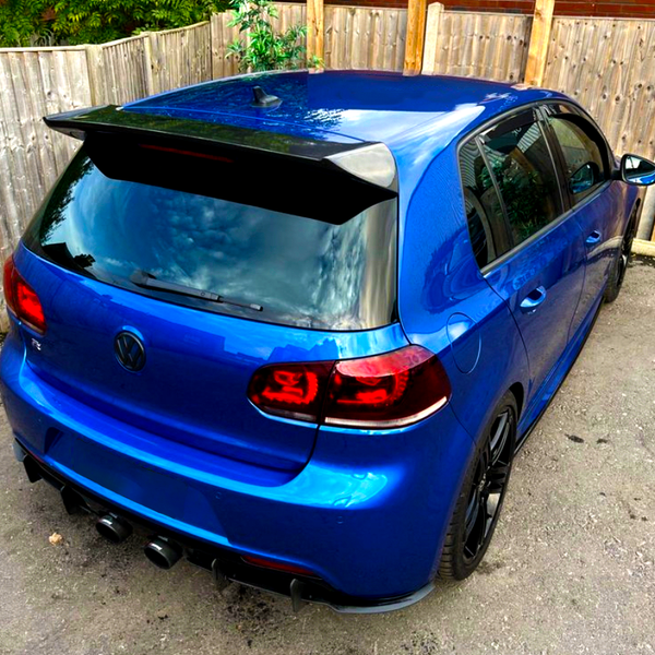 A blue hatchback car is parked on a pavement, featuring a spoiler and dual exhausts. It’s surrounded by wooden fencing and greenery. The Volkswagen logo is visible on the back.