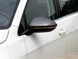 A car's side mirror with a carbon fiber finish is attached to the side of a white vehicle. The door handle and window are visible in an outdoor setting.