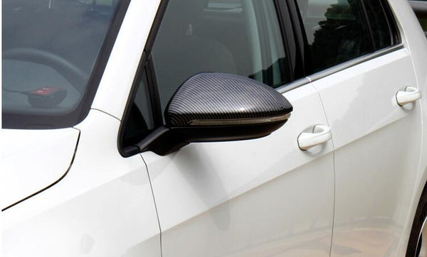 A car's side mirror with a carbon fiber finish is attached to the side of a white vehicle. The door handle and window are visible in an outdoor setting.