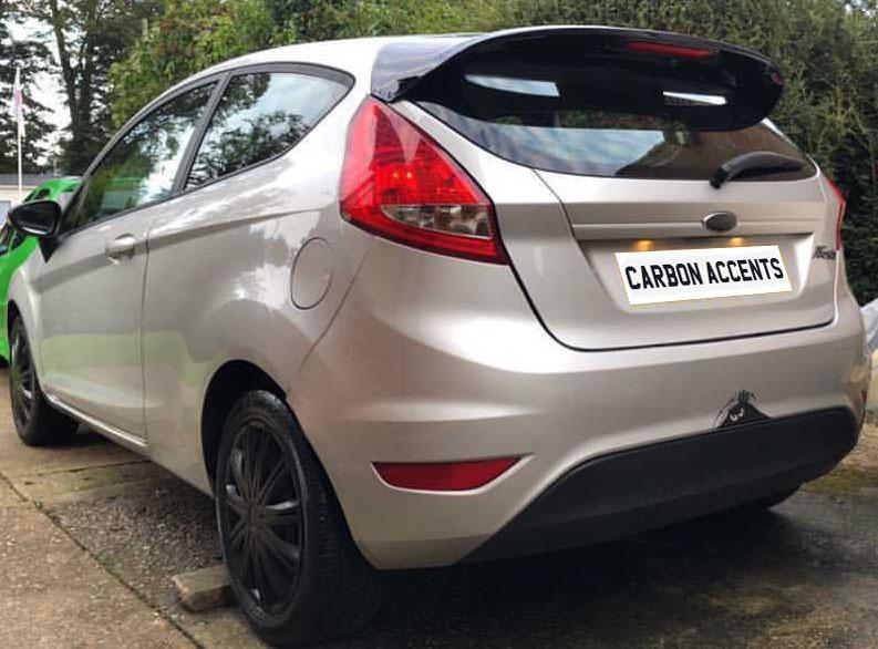 A silver hatchback car with tinted windows is parked on a driveway next to greenery. The license plate reads "CARBON ACCENTS." A small cartoon parachute is attached to the lower bumper.