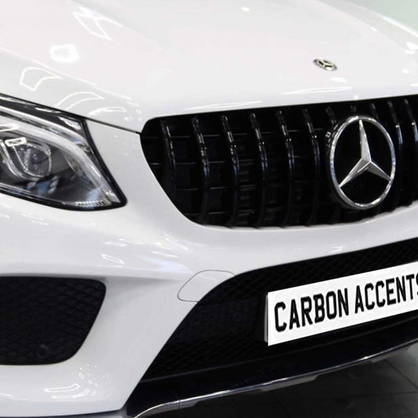 A white car with a prominent grille, featuring a Mercedes emblem, is parked indoors. The license plate reads "CARBON ACCENTS." The headlights are sleek and modern.