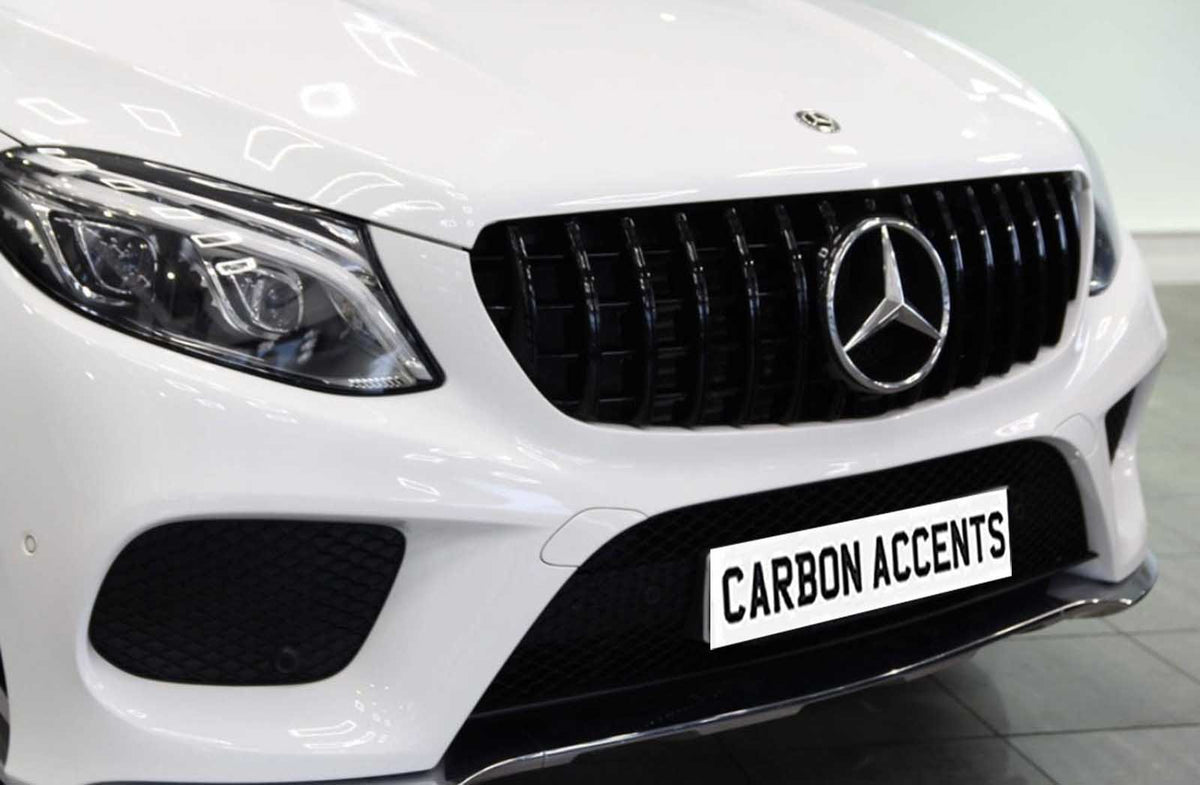 A white car with a prominent grille, featuring a Mercedes emblem, is parked indoors. The license plate reads "CARBON ACCENTS." The headlights are sleek and modern.