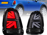 Two car taillights with a Union Jack design, one lit in red and the other unlit, positioned in front of a blue car on a road with a scenic background.