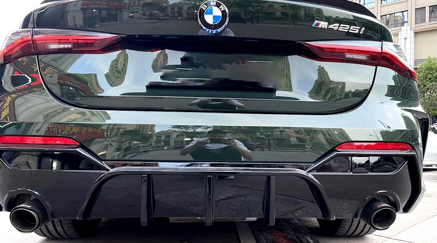 Car rear displays BMW logo and "M 425i" badge, with dual exhausts and diffuser. It is parked in an urban area with reflections of buildings on its shiny surface.
