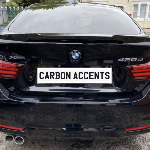 The black BMW 420d is parked, displaying "CARBON ACCENTS" on its license plate. It’s positioned in an outdoor setting with a blue car and a building nearby.