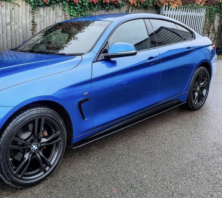 A blue sedan, parked on a wet road, features black alloy wheels and tinted windows. A wooden fence with climbing plants and a metal gate are in the background.