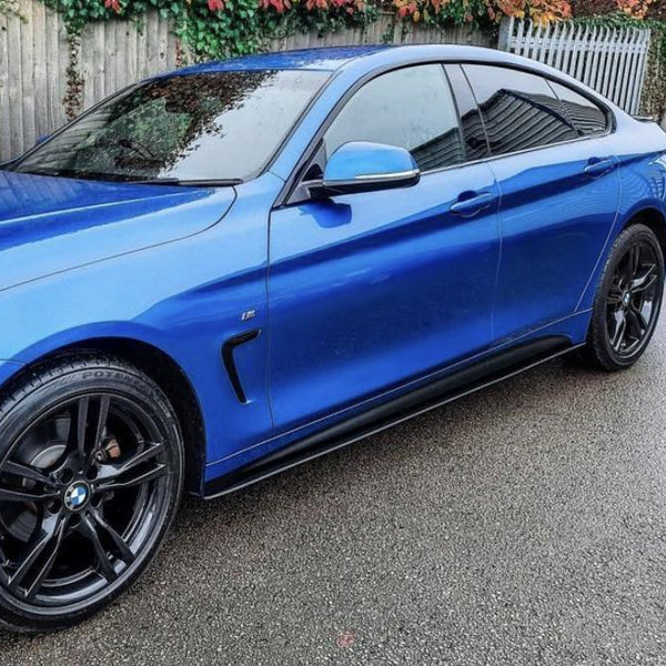 A blue sedan, parked on a wet road, features black alloy wheels and tinted windows. A wooden fence with climbing plants and a metal gate are in the background.