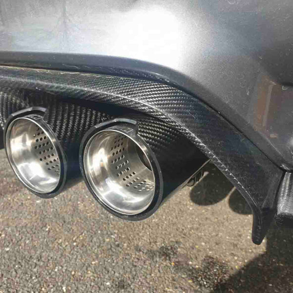 Two shiny metallic exhaust pipes are fixed onto a car's carbon fiber rear diffuser, with a textured road surface visible below in sunlight.