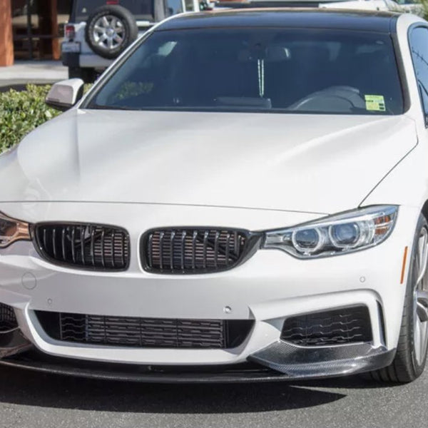 White car parked in a lot, with a sleek design featuring a prominent grille and headlights. Bushes and another vehicle are visible in the background under clear daylight.
