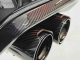 Dual exhaust pipes with a sleek carbon fiber finish emerge from a vehicle's rear bumper. The setting is a clean, bright environment, enhancing the contrast and details.