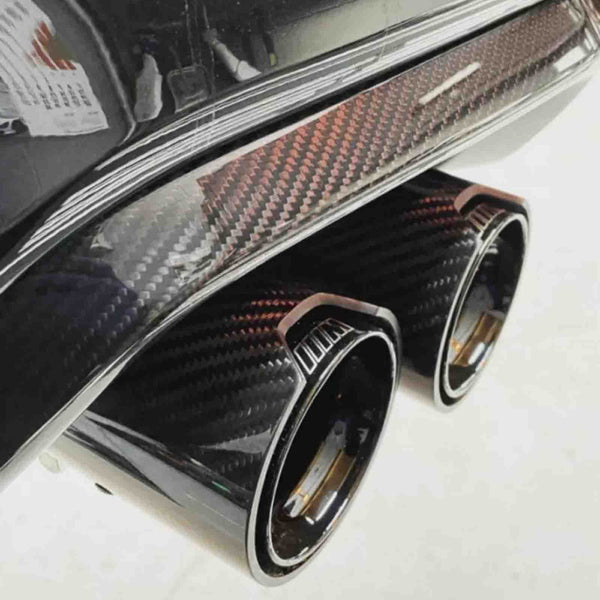 Dual exhaust pipes with a sleek carbon fiber finish emerge from a vehicle's rear bumper. The setting is a clean, bright environment, enhancing the contrast and details.