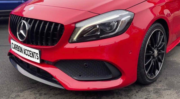 Red car with prominent front grille and sleek headlight, parked on a street. License plate reads "CARBON ACCENTS." A black tire and rim are visible in the foreground.