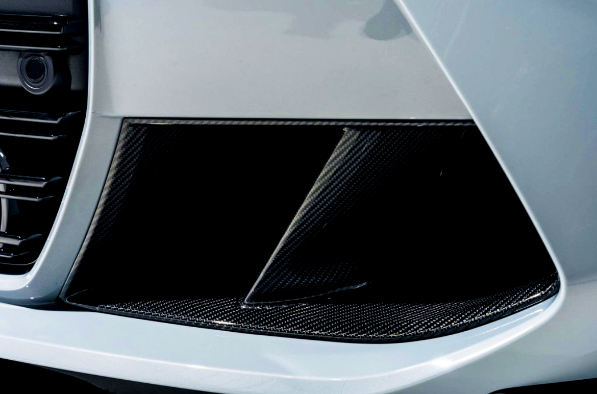 A close-up view of a car's front bumper area featuring a sleek, angular design with black carbon fiber components and a light-colored exterior, suggesting a sporty appearance.