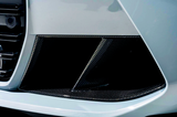 A close-up view of a car's front bumper area featuring a sleek, angular design with black carbon fiber components and a light-colored exterior, suggesting a sporty appearance.