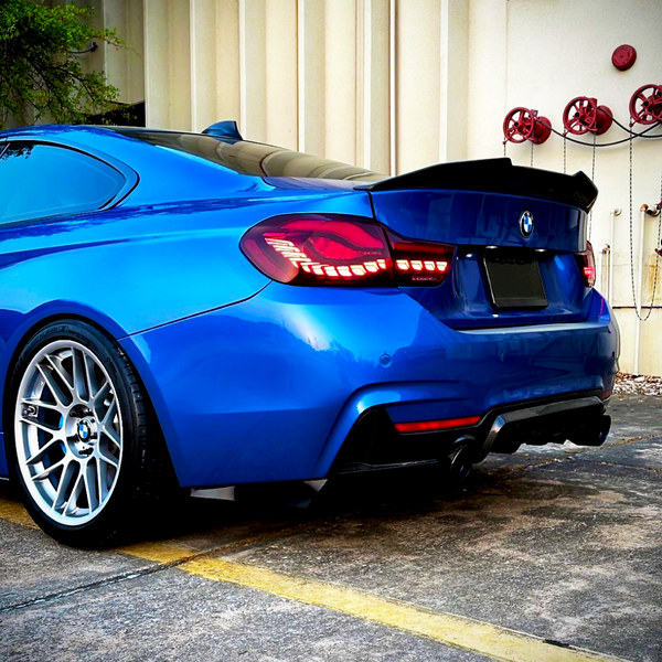 A blue sports car is parked on concrete, with tail lights illuminated. Red industrial valves are mounted on a beige wall in the background.