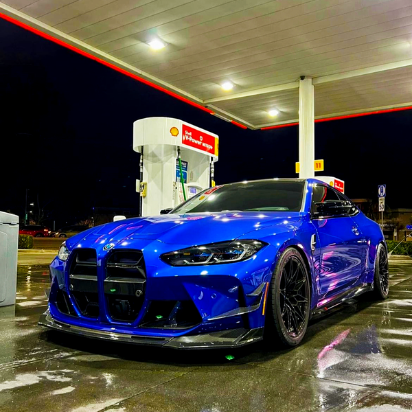 A blue sports car is parked at a gas station with a Shell V-Power Nitro+ pump, under bright canopy lights, reflecting on the wet pavement at night.