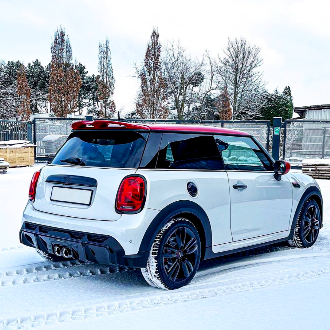 A white car with a red roof is parked on snow, leaving tire tracks. It's in a fenced area with trees and a building in the background.