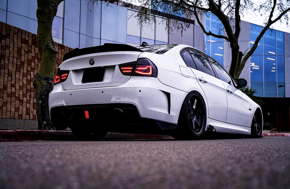White sports car parked, facing left, with illuminated tail lights. It is on a street with brick and glass buildings, and trees are nearby, creating an urban setting.