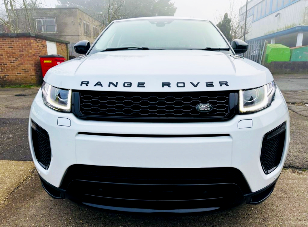 A white Range Rover, parked on a driveway, features a prominent front grille with the "Land Rover" logo. Overcast sky and industrial buildings set the backdrop.