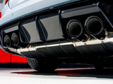 Exhaust pipes emit no smoke, under a sports car with a carbon fiber rear diffuser. The vehicle's exterior is glossy silver, set against a stark red and white garage wall.