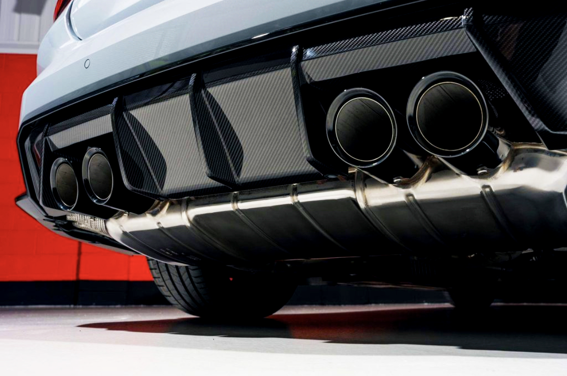 Exhaust pipes emit no smoke, under a sports car with a carbon fiber rear diffuser. The vehicle's exterior is glossy silver, set against a stark red and white garage wall.