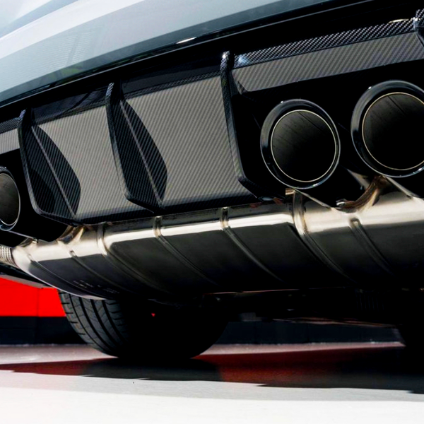 Exhaust pipes emit no smoke, under a sports car with a carbon fiber rear diffuser. The vehicle's exterior is glossy silver, set against a stark red and white garage wall.