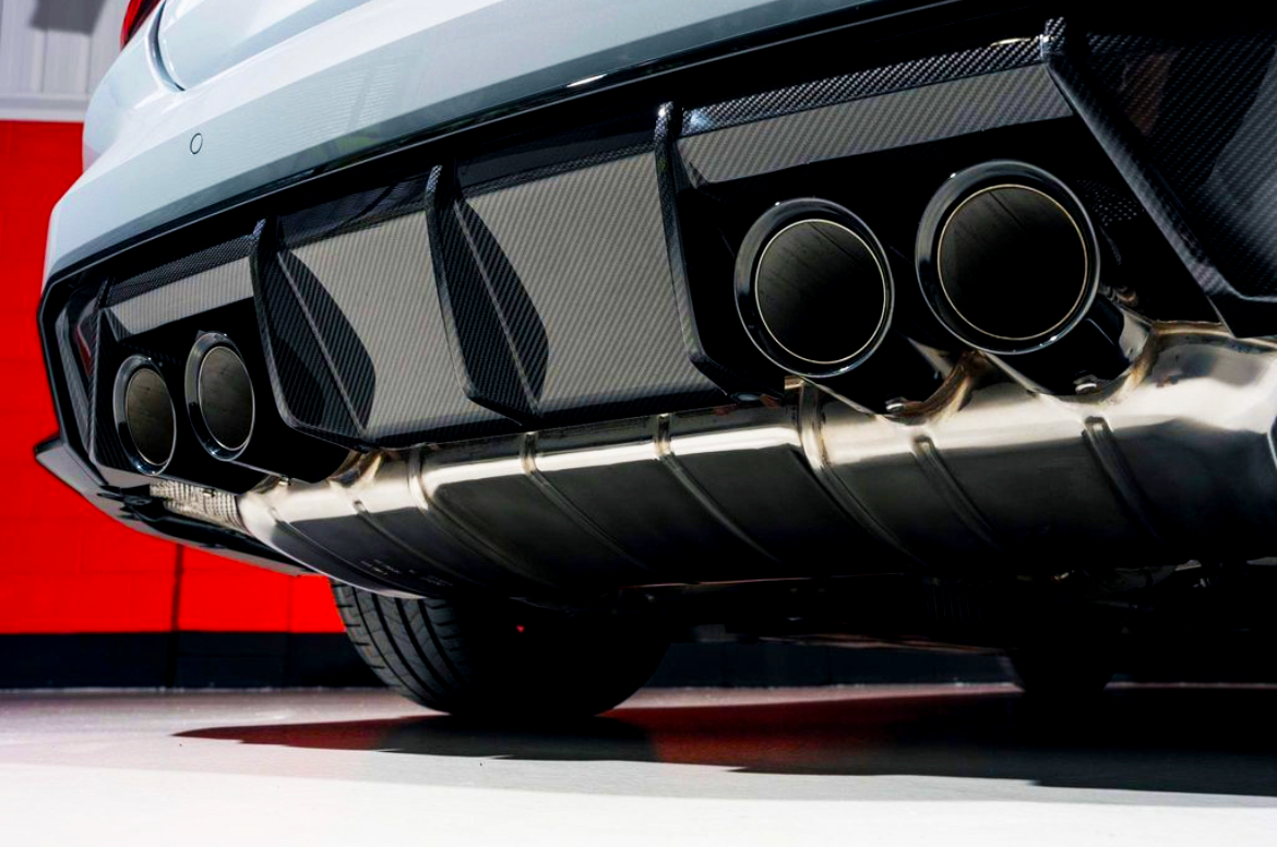 Exhaust pipes emit no smoke, under a sports car with a carbon fiber rear diffuser. The vehicle's exterior is glossy silver, set against a stark red and white garage wall.