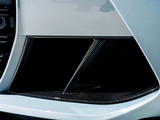 A car's carbon fiber air intake vent features a slatted design, positioned on the vehicle's front bumper. The glossy finish contrasts with the surrounding white bodywork, suggesting a sporty aesthetic.