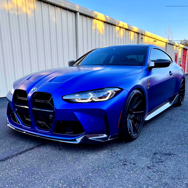 A blue sports car with sleek, angular design is parked on a paved surface. It is positioned in front of a beige industrial building under a clear blue sky.