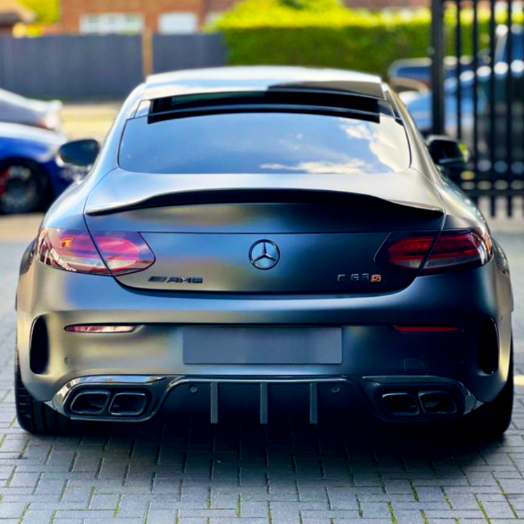 A matte black Mercedes-Benz AMG C63 parked on a paved surface, features visible exhausts and logos. Background includes parked cars and blurred greenery with a fence.