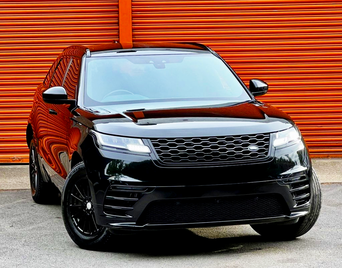 A black SUV is parked on a concrete surface, facing forward. Its sleek design reflects light, contrasting against the bright orange corrugated metal wall in the background.