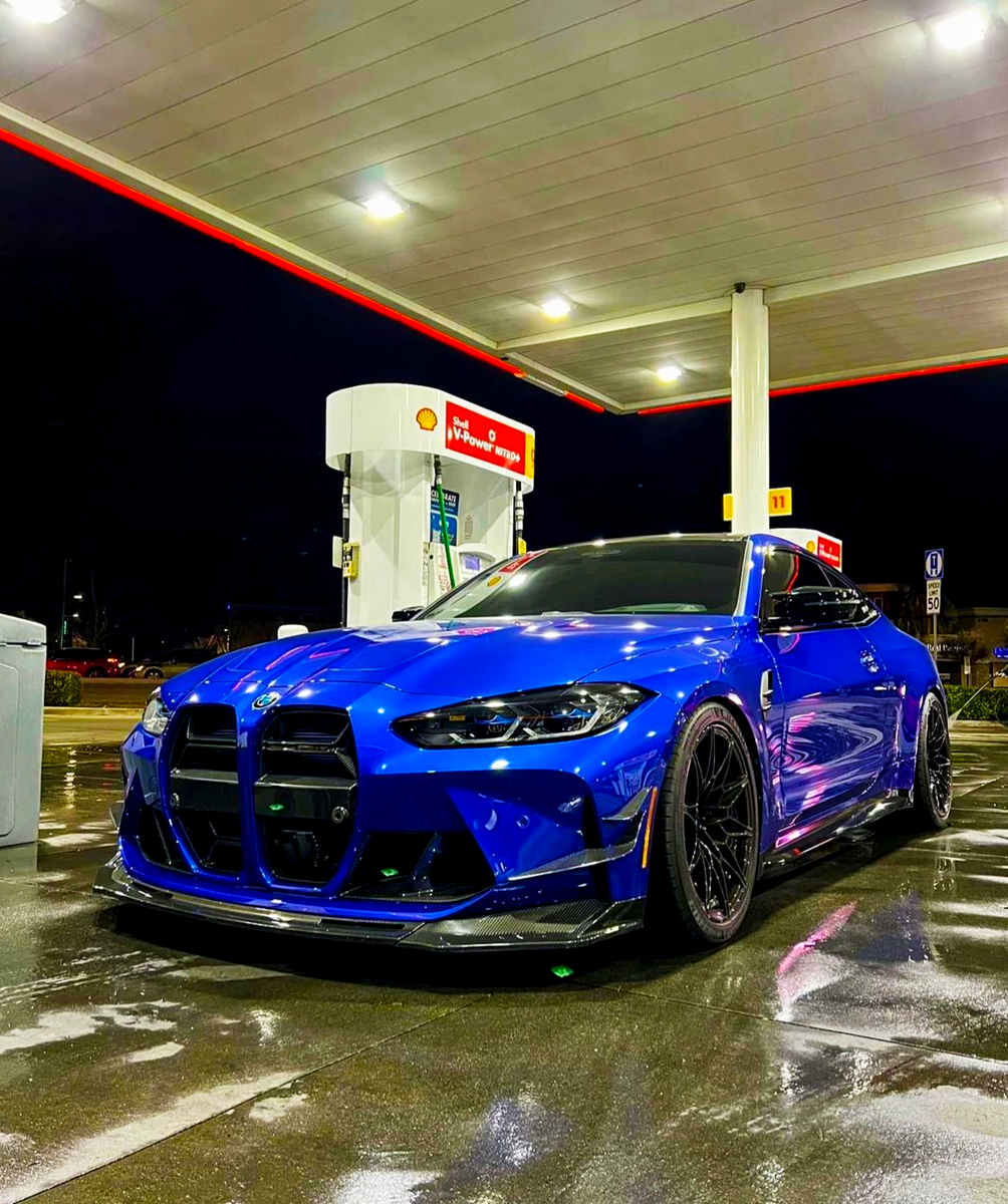 A shiny blue sports car is parked under a lit gas station canopy at night, reflecting the station lights on its wet surface.
