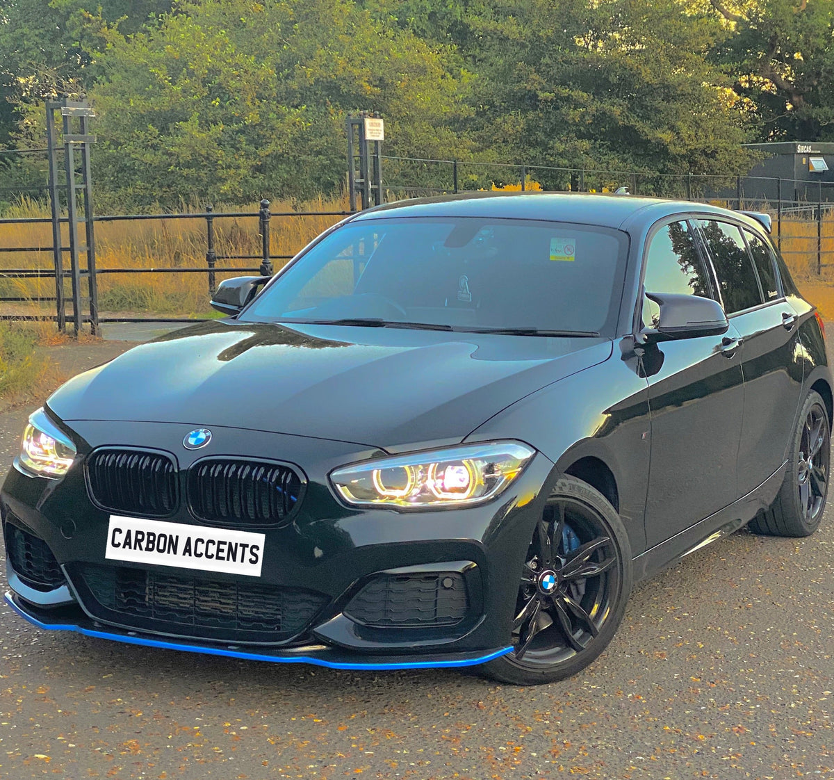 A black car with glowing headlights is parked on a road. It has a license plate reading "CARBON ACCENTS," surrounded by trees and metal railings in the background.