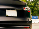 A sleek black car is parked outdoors, showing its rear with a blank license plate and visible taillights. A barrier and trees are in the background.